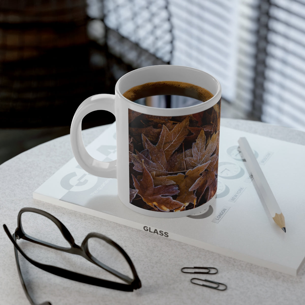 A large, white ceramic coffee mug is printed with an image of autumn leaves on the ground rimmed with a delicate frost.